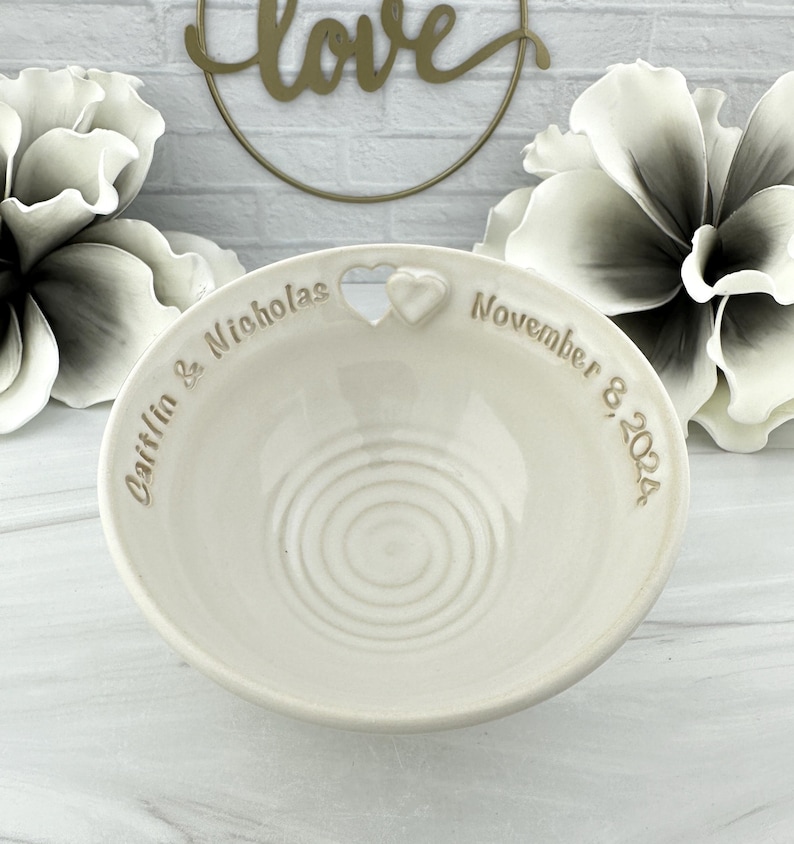 May include: White ceramic ring dish with a heart-shaped cutout and the inscription "Caitlin & Nicholas November 8, 2024".
