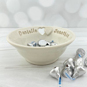 May include: White ceramic bowl with a heart-shaped cutout and the names "Danielle" and "Jonathan" inscribed on the rim. The bowl is filled with silver foil-wrapped chocolates.