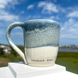 May include: A ceramic mug with a blue and white glaze. The top half of the mug is a deep blue, transitioning to a lighter blue and white. The words "Geenas mug" are printed on the lower portion of the mug.
