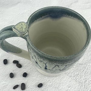 May include: A handmade ceramic mug with a blue and white glaze. The mug has a large handle and a heart-shaped detail on the handle. Several coffee beans are scattered around the mug.