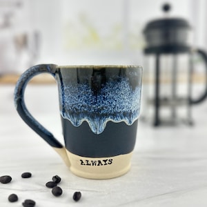 May include: A ceramic coffee mug with a dark blue and white speckled glaze. The mug has a black handle and the word "ALWAYS" printed on the base. Coffee beans are scattered around the mug.