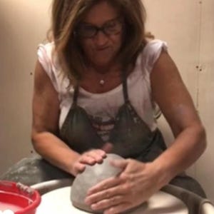 May include: A woman is working on a pottery wheel, shaping a gray clay pot with her hands.