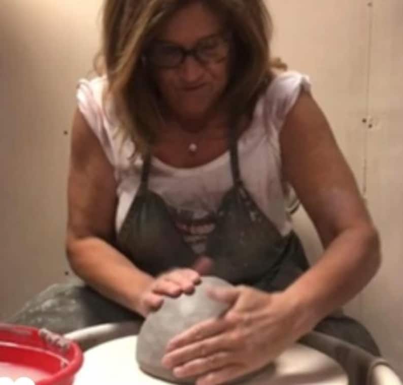 May include: A woman is working on a pottery wheel, shaping a gray clay pot with her hands.