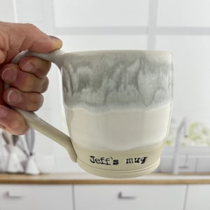 May include: A handmade ceramic mug with a light gray and white glaze. The mug has a large handle and the text "Jeff's mug" is printed on the base. The mug is held by a hand, showcasing its size and design.