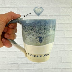 May include: A handmade ceramic mug with a blue and white glaze, featuring a curved handle and the inscription "Sabra's Mug." A small, heart-shaped ceramic accent hovers above the mug. The mug is held by a hand.