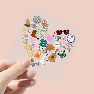 May include: A heart-shaped sticker with various colorful illustrations, including a disco ball, snake, boots, and guitar. Other elements include a lock, butterfly, and the number 13. The sticker is held by a hand against a pink background.