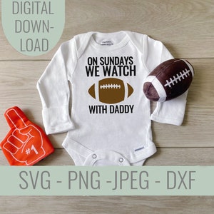 Pode incluir: Macacão branco de manga comprida com o texto "On Sundays We Watch With Daddy" e um gráfico de futebol americano marrom. Um dedo de espuma vermelho com o número um está no canto inferior esquerdo da imagem. Uma bola de futebol americano marrom está no canto superior direito da imagem.