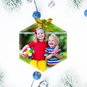 Op de afbeelding: Een kerstbal met een foto van twee kinderen, een meisje in een rood shirt en een jongen in een gestreept shirt, die lachen en een voetbal vasthouden. De bal is zeshoekig en heeft een gouden lint.