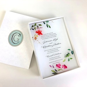 May include: A white wedding invitation with a floral design in shades of pink, red, and green. The invitation is in a white box with a light blue monogram. The text on the invitation reads: "Mr. and Mrs. Jay Butler, together with Mr. and Mrs. Ernest Pasarelli, request the pleasure of your company at the marriage of their children, Johanna Leigh and Christian Sekastian, Friday, the twenty-second of December, two thousand seventeen, at six o'clock in the evening, Coscaphe Ballroom, 132 North Second Street, Philadelphia, Pennsylvania, Dinner and dancing to follow, Black tie optional."