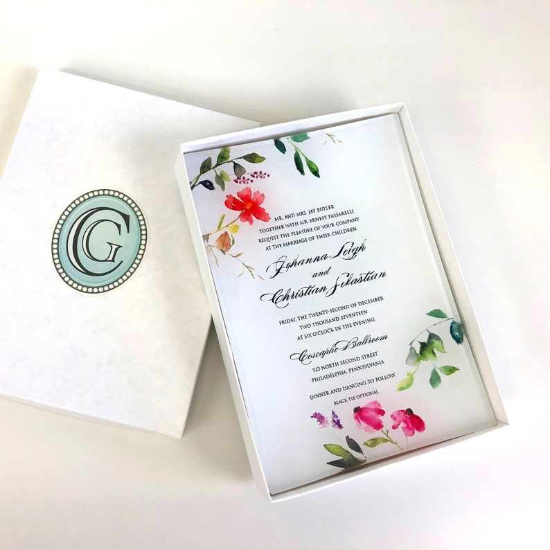 May include: A white wedding invitation with a floral design in shades of pink, red, and green. The invitation is in a white box with a light blue monogram. The text on the invitation reads: "Mr. and Mrs. Jay Butler, together with Mr. and Mrs. Ernest Pasarelli, request the pleasure of your company at the marriage of their children, Johanna Leigh and Christian Sekastian, Friday, the twenty-second of December, two thousand seventeen, at six o'clock in the evening, Coscaphe Ballroom, 132 North Second Street, Philadelphia, Pennsylvania, Dinner and dancing to follow, Black tie optional."
