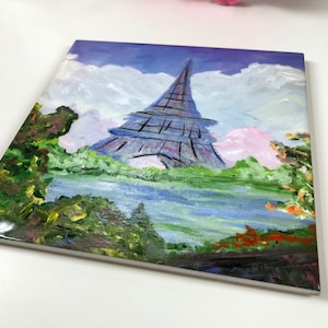 May include: A hand-painted ceramic tile featuring a colorful depiction of the Eiffel Tower in Paris, France. The tower is painted in shades of purple, blue, and black, with a green grassy area and a blue body of water in the foreground. The sky is painted in shades of blue and white with fluffy clouds.