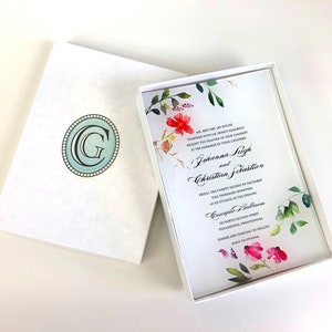 May include: A white wedding invitation with a watercolor floral design in shades of pink, red, and green. The invitation is in a white box with a light blue and white monogrammed "G" on the front. The text on the invitation reads: "Mr. and Mrs. Jill Butler, together with their parents, request the pleasure of your company at the marriage of their children, Johanna Leigh and Christina Jelastian, Friday, the twenty second of December, two thousand twenty, at six o'clock in the evening, at Coscgie Ballroom, 100 North Second Street, Philadelphia, Pennsylvania, Dinner and dancing to follow, Black tie optional."