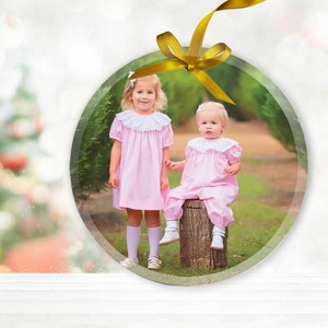 May include: Two young girls in pink dresses stand in front of a Christmas tree farm. One girl is holding the other's hand. The girls are smiling and looking at the camera. The image is framed in a round, clear ornament.