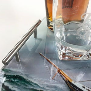 May include: A glass serving tray with a silver handle. The tray has a printed image of a sailboat on a stormy sea. A glass with the word "First" etched on it is on the tray.