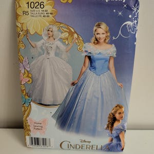 May include: Simplicity 1026 Disney Cinderella home sewing pattern envelope featuring two dress variations. One dress is white with a high collar, and the other is a light blue off-the-shoulder gown with a full skirt. Includes size U.S. 14-22.