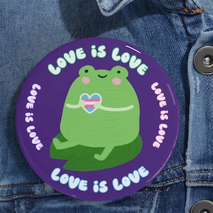 May include: A circular, purple badge featuring the text "LOVE IS LOVE" around a green frog holding a heart in the colours of the transgender pride flag. The frog is sat on a green leaf.