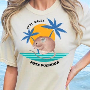 May include: Cream-colored t-shirt featuring a graphic of a mouse, palm trees, and a sun. The text on the shirt reads "Stay Salty" above the mouse and "Pots Warrior" below. The shirt is worn near a beach.