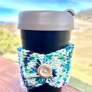 May include: A black travel mug with a gray lid and a colorful knitted cozy. The cozy is white with blue, green, and purple flecks, secured with a wooden button that reads "Esdon Lodge Farm".