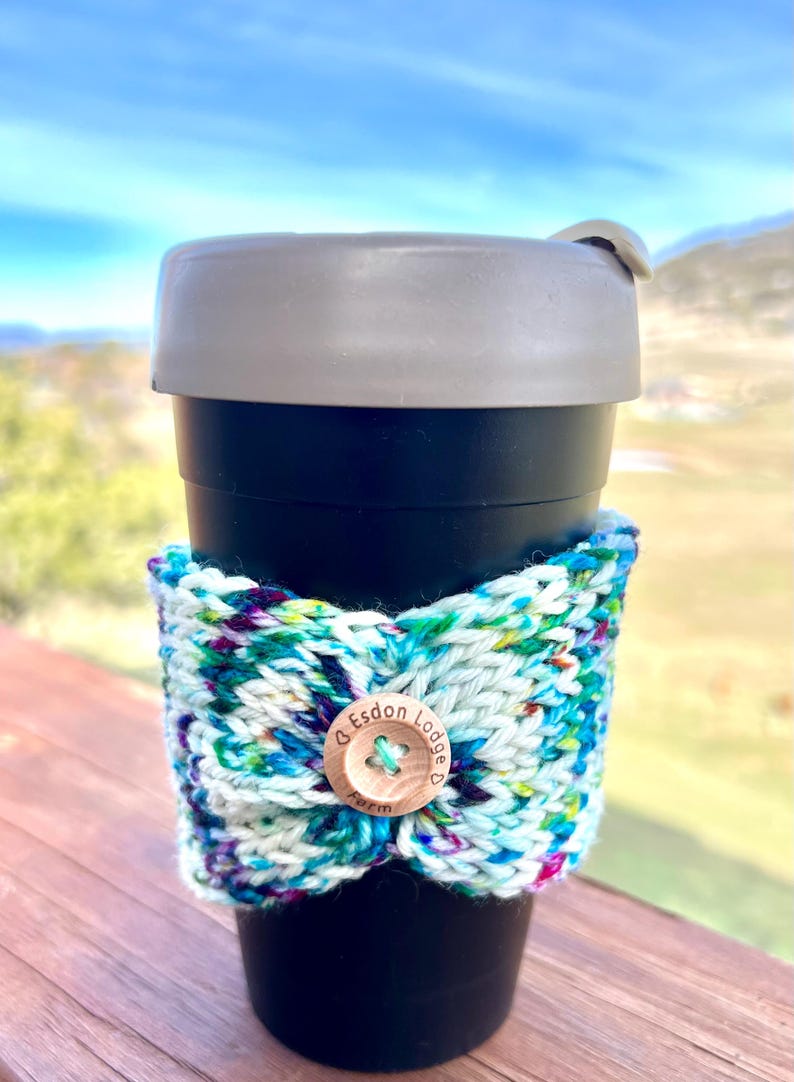 May include: A black travel mug with a gray lid and a colorful knitted cozy. The cozy is white with blue, green, and purple flecks, secured with a wooden button that reads "Esdon Lodge Farm".