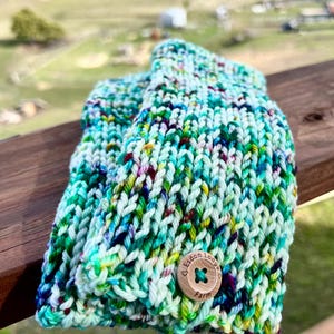 May include: Hand-knitted mittens in shades of teal, green, and purple with a wooden button closure. The mittens are made of thick yarn and have a textured appearance. The button is engraved with the text "Esdon Lodge Farm".