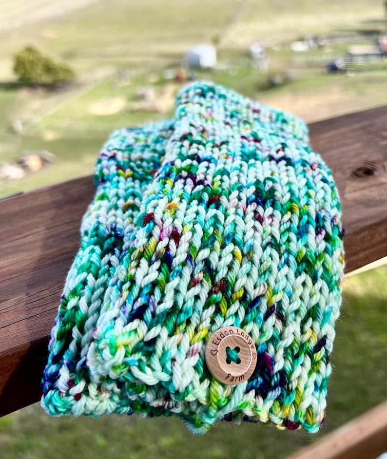 May include: Hand-knitted mittens in shades of teal, green, and purple with a wooden button closure. The mittens are made of thick yarn and have a textured appearance. The button is engraved with the text "Esdon Lodge Farm".