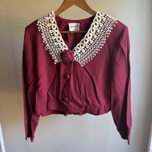 May include: A deep burgundy long-sleeve jacket featuring a wide, cream-colored crocheted lace collar, a fabric-covered rose accent at the neckline, and two matching fabric-covered buttons down the front. The interior label reads Jeffrey & Dara.