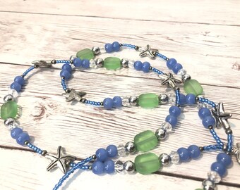 May include: A beaded necklace featuring blue, green, and silver beads, interspersed with silver starfish charms. The necklace is strung on a blue cord, creating a nautical theme. The beads are various shapes and sizes.