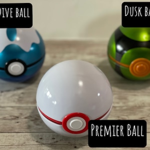 May include: Three colorful Pok&eacute; Balls: a blue and white Dive Ball, a white Premier Ball with red accents, and a green and black Dusk Ball. Each ball has a white button in the center. The text "DIVE BALL", "PREMIER BALL", and "DUSK BALL" are displayed.