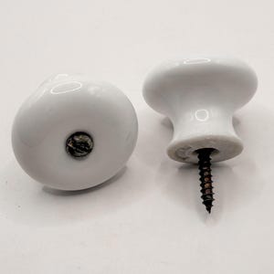 May include: Two white ceramic cabinet knobs with a rounded shape. One knob is lying on its side, while the other stands upright, revealing a black screw. The knobs are set against a white background. The text "Sweet Cottonwood" is at the bottom.