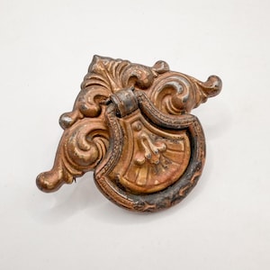 May include: Ornate antique bronze drawer pull with a shield-like design and scrollwork. The hardware has a weathered appearance, with shades of brown and copper. The pull is likely for a cabinet or drawer, adding a vintage touch.
