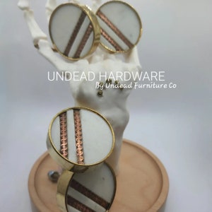 May include: Three round white and gold cabinet knobs with copper accents. The knobs are displayed on a white hand and a wooden surface. The text "UNDEAD HARDWARE by Undead Furniture Co." is visible in the image.