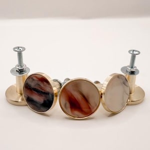 May include: Five decorative cabinet knobs with brass hardware. The knobs have a circular design with a marbled appearance in shades of red, brown, and cream. The knobs are attached to silver screws.