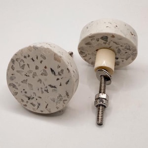 May include: Two round cabinet knobs with a speckled, light gray and white terrazzo design. One knob is attached to a screw with a nut and washer. The other knob is positioned to the left. The knobs are for furniture and home decor.