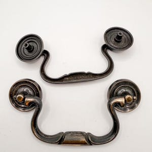 May include: Two antique bronze-colored drawer pulls with a curved handle design. Each pull has a circular backplate and a decorative handle. The pulls are displayed against a white background. The text "SWEET COTTONWOOD" is visible at the bottom of the image.