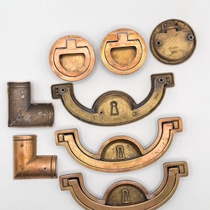 May include: An assortment of antique brass hardware, including drawer pulls, handles, and corner brackets. The hardware features a variety of shapes and sizes, with a warm, aged patina. The pieces are arranged on a white surface.