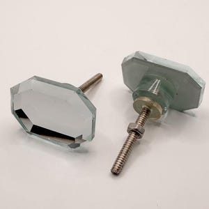 May include: Two clear, faceted glass cabinet knobs with silver-colored threaded posts and nuts. The knobs have an octagonal shape and are designed for furniture or cabinetry. The background is a plain white surface.
