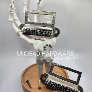 May include: Two silver drawer pulls with the word "Apothecary" engraved on them. The pulls are on a wooden surface with screws.