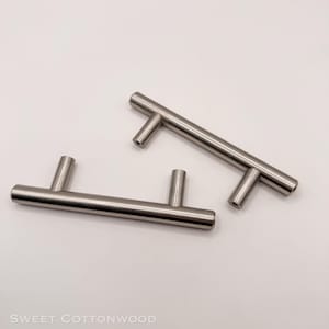 May include: Two stainless steel cabinet pulls with a T-bar design. The pulls are silver and have a brushed finish. The handles are designed for easy gripping and are suitable for drawers and cabinets.