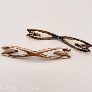 May include: Two vintage metal drawer pulls with a unique arrow-like design. One is a bronze color, and the other is a darker, aged metal. The pulls have a distinctive shape, with a central opening and mounting holes.