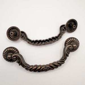May include: Two antique bronze-colored drawer pulls with a decorative, ornate design. Each pull features a circular backplate and a curved handle with a twisted rope-like pattern. The pulls are likely for furniture restoration.