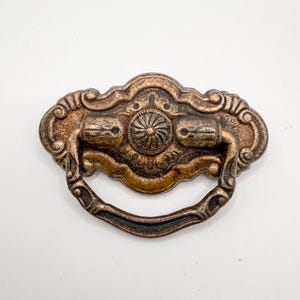 May include: Ornate bronze-colored drawer pull with a decorative design. The pull features a central floral motif and a curved handle. The metal hardware has an aged, antique appearance, perfect for vintage furniture restoration projects.