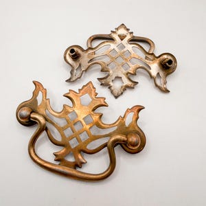 May include: Two ornate, vintage-style drawer pulls. One is a tarnished silver color, and the other is a warm, aged brass. Both feature a decorative openwork design with a handle and mounting hardware.