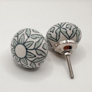 May include: Two ceramic cabinet knobs with a floral design in white and light green. One knob is shown with the screw and metal base attached. The knobs are round and have a glossy finish. The text "SWEET COTTONWOOD" is at the bottom.