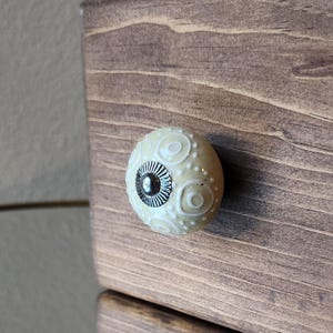 May include: Close-up of a wooden drawer with a decorative knob. The knob is off-white with a pattern of white swirls and a dark center. The drawer is stained a medium brown color.