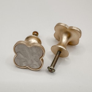 May include: Two gold-colored clover-shaped cabinet knobs. One knob has a white and gray marbled center. The other knob is lying on its side, showing the screw. The text "SWEET COTTONWOOD" is at the bottom of the image.
