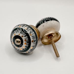May include: Two decorative ceramic cabinet knobs. The knobs are white with black and gold accents. One knob is in focus, displaying a detailed pattern. The other knob is slightly out of focus, showing the screw and mounting hardware. The knobs are on a white background.