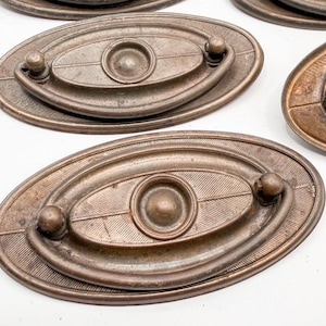 May include: Antique bronze oval drawer pulls with a central circular design and a curved handle. The pulls have a textured surface with a leaf-like pattern. The hardware is likely for furniture restoration.