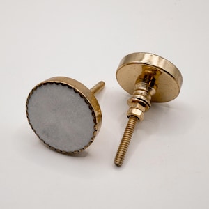 May include: Two decorative cabinet knobs. Each knob features a round, light gray center with a gold-colored, scalloped edge. The knobs have a gold-colored base and screw. The text "SWEET COTTONWOOD" is at the bottom of the image.