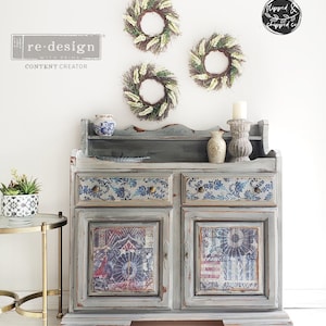 May include: A gray painted wooden cabinet with two drawers and two doors. The doors and drawers have a blue and white floral pattern. The cabinet has a distressed finish and is decorated with three wreaths of white flowers.