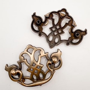 May include: Two antique bronze-colored drawer pulls with ornate, openwork designs. Each pull features a curved handle and decorative backplate with intricate patterns. The pulls are likely for a cabinet or dresser.